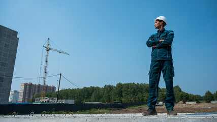 A builder in work clothes and a helmet stands on a construction site against the background of a construction crane. 