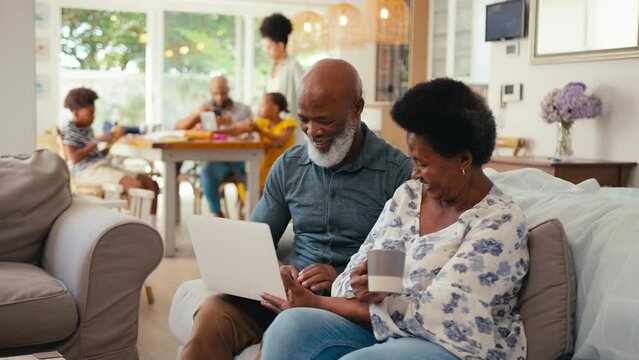 Senior couple at home looking at laptop together with multi-generation family in background - shot in slow motion