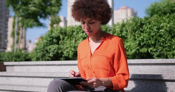 Happy young African American woman using digital tablet pad sufring internet inspired with new ideas sitting outdoors