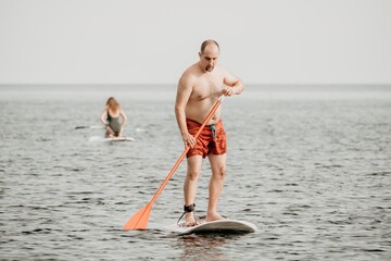 Active mature male paddler with his paddleboard and paddle on a sea at summer. Happy senior man stands with a SUP board. Stand up paddle boarding - outdor active recreation in nature.