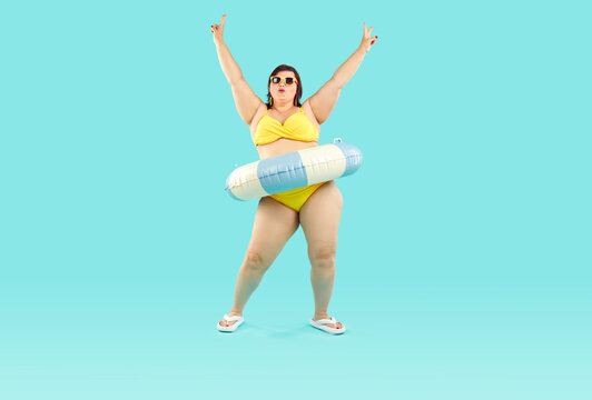 Happy confident fat woman having fun at beach party on summer vacation holiday. Plus size lady in yellow bikini, with inflatable rubber floater around waist dancing isolated on blue background