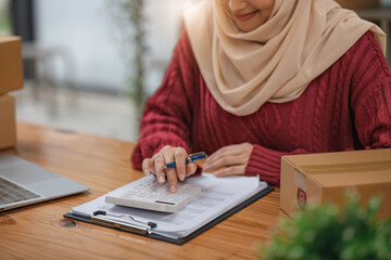 A beautiful and happy young Asian Muslim female online seller using a calculator, checking products stocks and calculating sales and income at her desk.