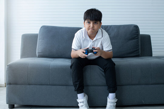 Concentrated Indian boy excited playing video game, sitting on couch and holding controller having fun in gamer emotion, kid gaming addiction concept