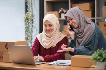 A happy young Asian Muslim female online seller working with her employee in the office. SME business, online shop owner