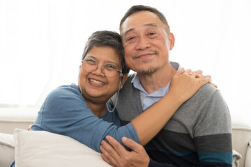 Happy smiling Asian elderly couple hugging together at home. Asian senior man embracing elderly woman on sofa in living room at home. Retirement, health care, relax and spending time concept