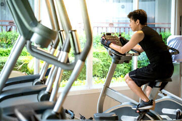 Active Asian sport man in black sportswear cycling on an upright stationary bike or upright exercise bicycle. Indoor cardio training workout and burn calories in fitness gym © Summer Paradive