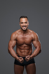 Fototapeta na wymiar Breathtakingly handsome male model with a chiseled physique flashes a charming smile as he poses in black briefs against grey background, emanating charisma and sex appeal