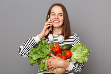 Beautiful happy joyful woman embracing fresh vegetables returns from supermarket wearing striped shirt isolated over gray background talking on mobile phone.