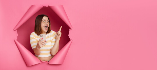 Surprised happy woman wearing striped shirt breaking through pink paper hole holding samrtphone pointing away at copy space for advertisement or promotional text.