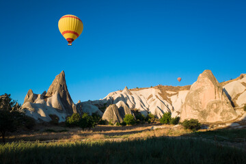 A yellow hot air balloon flies against a clear blue sky over the rock formations of Fairy Chimneys...