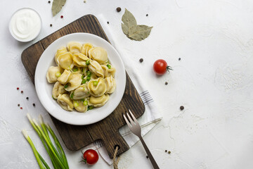 Dumplings boiled in chicken broth and sprinkled with green onions on a white plate. Cherry tomatoes, peppercorns and bay leaves on a light table. Sour cream in a white bowl in the background. Top view