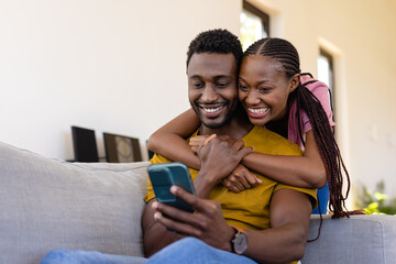 Happy african american couple embracing and using smartphone in living room