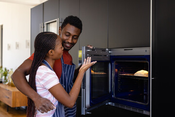 Happy african american couple in aprons baking bread using oven in kitchen