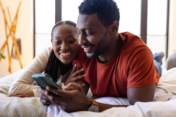 Happy african american couple using smartphone lying on bed in bedroom