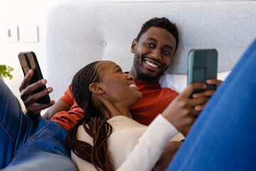 Happy african american couple using smartphones lying on bed in bedroom