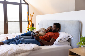 African american couple using tablet lying on bed in bedroom