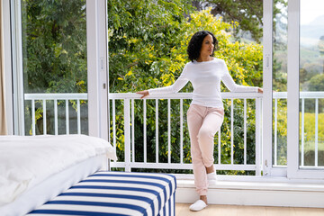 Happy biracial woman in slippers relaxing at balcony in bedroom with treetops in background