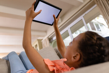 Happy biracial girl lying on couch using tablet with copy space on screen in living room