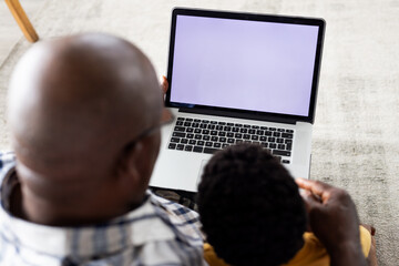 African american grandfather and grandson sitting on sofa, using laptop with copy space on screen