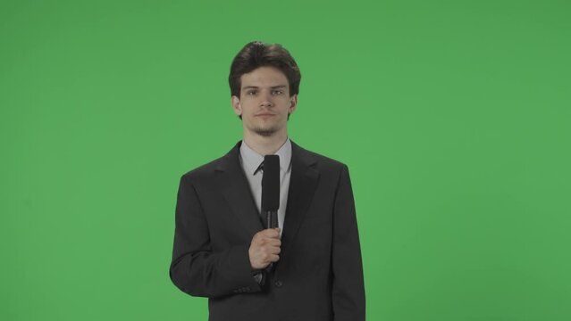 Young journalist with a microphone in front of a green screen. A man in a suit speaks and points to a green screen. TV news concept. Advertising area, workspace mock up. HDR BT2020 HLG Material.