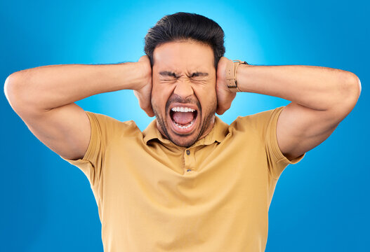 Man covering his ears while screaming in a studio for angry, upset or mad argument expression. Crazy, shout and young male person with an open mouth for loud voice gesture isolated by blue background