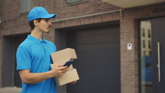 Male Courier Going Along Building Holding the Paper Boxes and Mobile Phone in Hand. Delivery Man Looking for Delivery Address on Street Using Smartphone While Delivering Outdoors