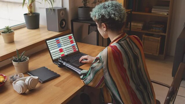 Back view of young Caucasian woman with short curly grey blue hair and prosthetic arm working on laptop computer from home office