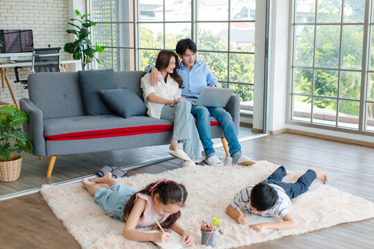 Asian happy cheerful joyful family husband and wife lover couple sitting on carpet floor helping teaching little boy son and girl daughter painting drawing cartoon with color pencils in living room