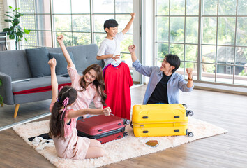 Fototapeta premium Asian little boy and girl helping father and mother preparing packing clothes and stuff into trolley luggages on living room carpet floor for traveling on holiday weekend vacation family road trip