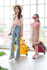 Portrait shot of Asian happy family mom and daughter wearing sunglasses and hat standing with red and yellow trolley luggages look at camera waiting for departure time in airport hallway for vacation