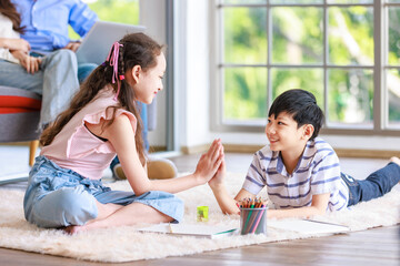Asian happy cheerful joyful little brother and sister sitting laying lying down on carpet floor high five together while painting drawing cartoon with color pencils together in living room at home