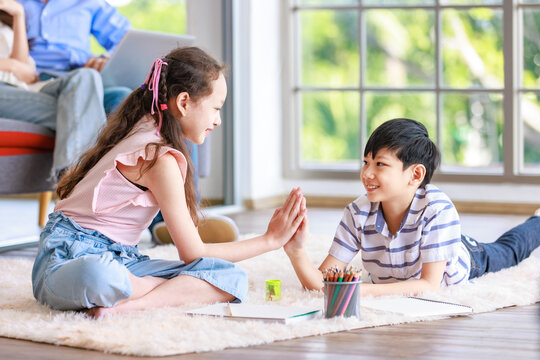 Asian happy cheerful joyful little brother and sister sitting laying lying down on carpet floor high five together while painting drawing cartoon with color pencils together in living room at home