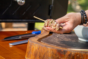 Obraz premium A white man's hand holding a chicken heart stick next to some knives and a white bowl above a rustic wooden board ready to bring it to the barbecue grill for dinner or lunch