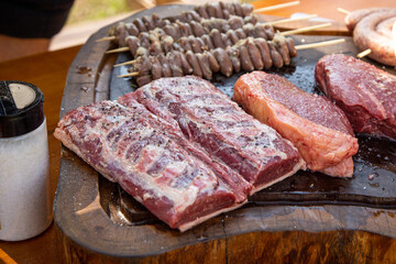 A delicious raw mix of meat composed by chicken wings, sausage, lamb loin and picanha medallion resting on a rustic wooden board next to a pot of salt ready for a gaucho traditional barbecue lunch