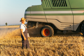 An elderly farmer with a digital tablet in his hands walks through a field of wheat under harvest,...