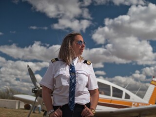 Blonde female pilot dressed in uniform posing near an airplane.