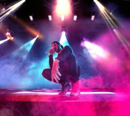 Artistic young man in stylish clothes with dreads performing on stage at nightclub, singing rap over multicolor neon light. Concept of music, performance, art, talent, nightlife, joy, party, lifestyle - obrazy, fototapety, plakaty