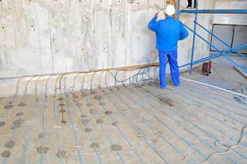 A man wearing a blue uniform is seen installing an underfloor heating cable on a concrete surface,...