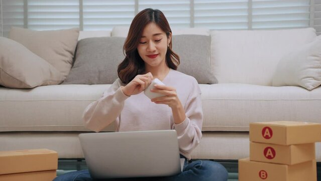 Young asian woman selling online receiving order via laptop and using label printer to printing customer address attach to the parcel box. Business Online and delivery shop concept