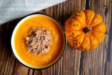 on a brown wooden table there is a gray round plate of pumpkin cream soup with tuna next to a small pumpkin.  gluten-free, vegan.  diet.  view from above.  menu calendar