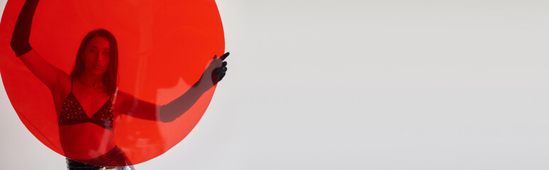 fashion photography, latex style, young asian woman with brunette hair posing in bra and gloves and holding red round shaped glass on grey background, fashion choices, behind glass, banner