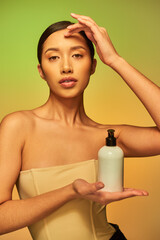 Obraz na płótnie Canvas product presentation, young asian woman with bare shoulders holding cosmetic bottle with beauty product and looking at camera on green background, brunette hair, glowing skin