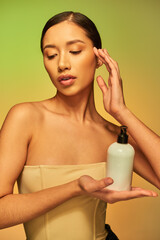 Obraz na płótnie Canvas product presentation, beauty product, young asian woman with bare shoulders holding cosmetic bottle with body lotion and posing on green background, brunette hair, glowing skin