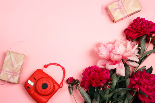 Modern instant camera with peony flowers and gift boxes on pastel pink background.