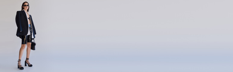 Fototapeta premium trendy look, fashion forward, brunette asian woman in sunglasses posing with feathered purse on grey background, model in latex shorts, bra, black jacket and gloves, youth, full length, banner