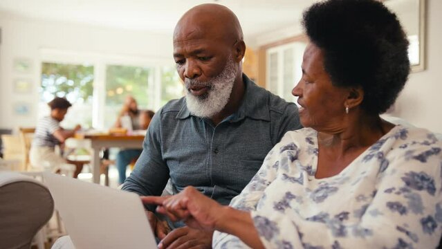 Senior couple at home looking at laptop together hugging after booking holiday or making major purchase with multi-generation family in background - shot in slow motion