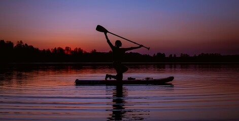 Female figure holding a paddle standing on a board at sunset.