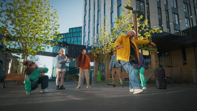 Young And Athletic B-Boy Breakdancing To Break Beats On The City Street Among Modern Buildings In The Circle Of Friends. Fashionable Young People Supporting Dancer, Live Streaming Via Smartphone.