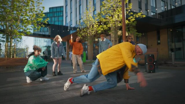 Young And Athletic B-Boy Breakdancing To Break Beats Downtown On City Street Among Modern Buildings In The Circle Of Friends. Stylish Young People Supporting Dancer, Live Streaming Via Smartphone.