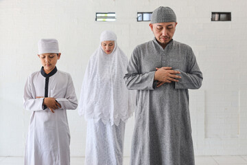 Religious Asian muslim family praying together, stands with hand in chest are doing salat in mosque